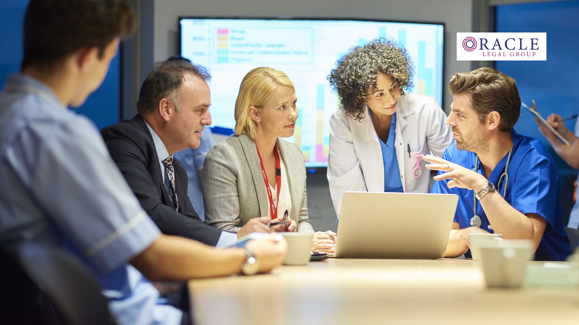 A diverse group of healthcare professionals and administrators gathered around a laptop during a training session, with performance charts on a screen; Oracle Legal Group logo appears in the corner